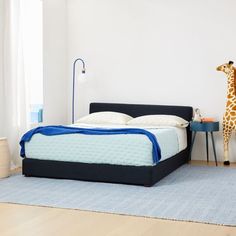 a giraffe standing next to a bed in a room with blue rugs