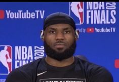 a man with a beard wearing a black shirt and beanie sitting in front of a blue wall