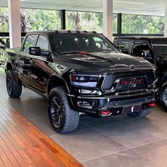 two black trucks are parked in a showroom with wood flooring and large windows