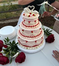 a wedding cake is sitting on a table with red roses and two people in the background