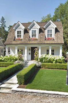 a white house with lots of flowers in the front yard and bushes on either side