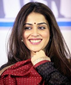 a smiling woman with long hair wearing a red and black top, standing in front of a wall