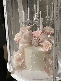 a white cake with pink flowers and candles in a glass case on top of a table