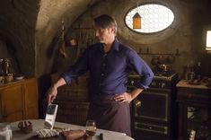 a man cutting food on top of a wooden table in a kitchen next to a round window