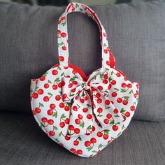 a red and white bag with cherries on it sitting on a gray couch next to a pillow