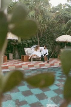 a man and woman sitting on a bench next to a pool with an umbrella in the background