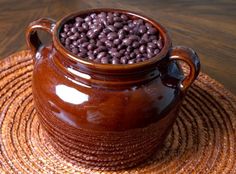 a brown pot filled with beans sitting on top of a straw place mat next to a wooden table