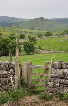 a stone wall and gate in the middle of a grassy field with mountains in the background