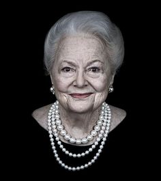 an old woman with pearls on her neck and necklace, smiling at the camera in front of a black background