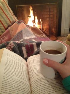 a person holding a cup of coffee while reading a book in front of a fire place