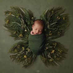 a newborn baby is wrapped in a green blanket surrounded by grass and flowers on a gray background