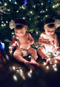 two babies sitting next to each other in front of a christmas tree with lights on it