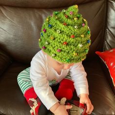 a small child wearing a green crocheted christmas tree hat sitting on a couch