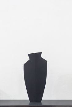 a black vase sitting on top of a wooden table next to a white wall in front of it