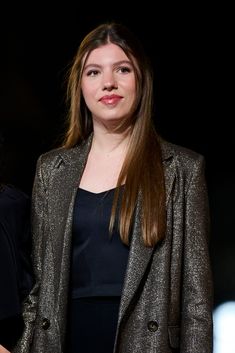 a woman with long hair standing in front of a black background wearing a silver blazer