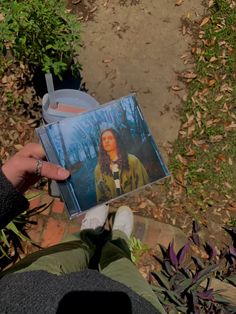 someone is holding up a cd in their hand while sitting on the ground near some plants