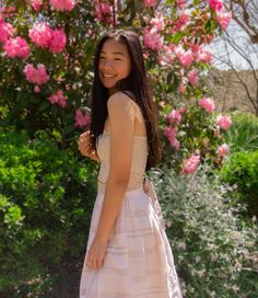 a woman in a white dress standing next to some pink flowers and smiling at the camera