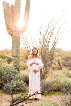 a pregnant woman standing in front of a cactus with her arms around her stomach and wearing a white dress