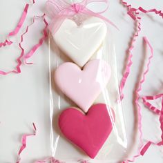 three heart shaped cookies in plastic bags on a white surface with pink streamers around them