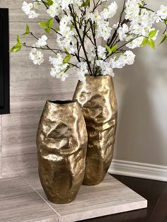 two gold vases with flowers in them sitting on a marble table next to a wall