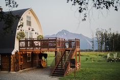 a wooden cabin with stairs leading up to the front door, and a large white barn in the background