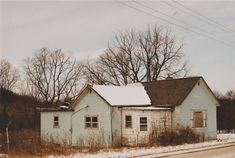 an old run down white house with snow on the roof and trees in the background