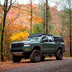 a green truck parked on the side of a road in front of trees and leaves