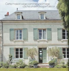 a large white building with green shutters on the front and side windows, surrounded by greenery