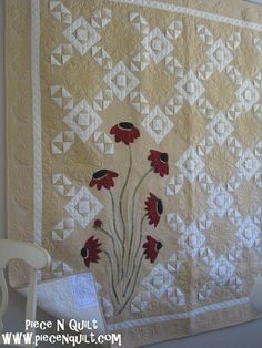 a quilted wall hanging with red flowers on it and a white chair next to it