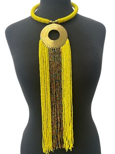 This stunning necklace brings together the beauty of African culture with elegance. The brass pendant features intricate engraving and a long fringe of yellow beaded strands, creating a unique and eye-catching piece. The necklace is perfect for any occasion and will add a touch of sophistication to any outfit. The beaded strands are crafted from high-quality materials, ensuring durability and longevity. This necklace is a must-have for anyone who loves African art and jewellery. Material: Beads and decorative brass metal. Fringe - 40cm Neck Width - 50cm Colour: Yellow, Gold, Multicolour Weight: 200g Gold Jewelry With Beaded Fringe For Festival, Unique Yellow Beaded Necklaces For Festival, Bohemian Gold Lariat Necklace With Dangling Beads, Gold Bohemian Lariat Necklace With Dangling Beads, Unique Yellow Beaded Necklace For Festivals, Handmade Adjustable Gold Tassel Necklace, Adjustable Yellow Lariat Necklace, Traditional Yellow Beaded Festival Necklaces, Traditional Yellow Beaded Necklaces For Festivals