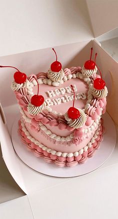 a pink heart shaped cake with cherries on top in a white cardboard box next to a knife and fork