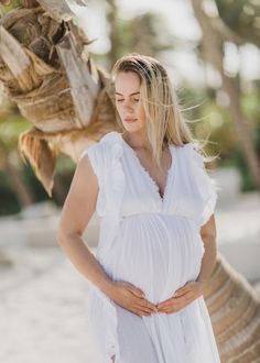 a pregnant woman wearing a white dress standing on the beach with her hands in her stomach