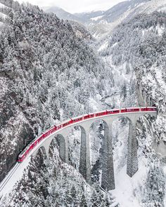 a red train traveling over a bridge in the middle of snow covered mountains and trees