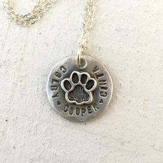 Pet memorial necklace This cat paw or dog paw necklace is a sweet reminder of your favorite pets. It also makes a lovely pet memorial necklace. The necklace can hold up to 22 characters/spaces combined. I can separate names with a tiny heart or a space.The only font I can use is the one shown in the photos and is available only in ALL UPPERCASE. The charm measures about 20mm across and the disc has hammered pattern. The photo shows it finished with a light matte/brush finish. But you can also op Small Personalized Sterling Silver Necklace, Hypoallergenic Sterling Silver Dog Tag Jewelry, Adjustable Paw Print Jewelry For Anniversary, Round Pendant Necklace With Paw Print For Gift, Nickel-free Dog Tag Jewelry For Personalized Gifts, Personalized Hand Stamped Dog Tag Jewelry, Sterling Silver Paw Print Necklace For Gift, Sterling Silver Paw Print Jewelry For Gifts, Silver Paw Print Jewelry As Gift