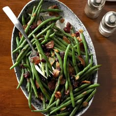 a bowl filled with green beans and bacon on top of a wooden table next to two salt shakers