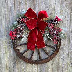 a wagon wheel decorated with red velvet bows and pineconis is hung on the side of a wooden fence