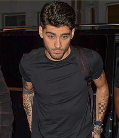 a man in black shirt and jeans with tattoos on his arm walking towards a car