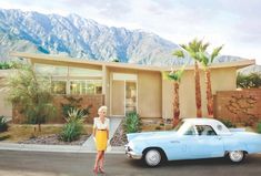 a woman standing next to a blue car in front of a house with mountains in the background