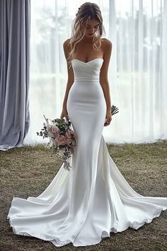 a woman standing in front of a window wearing a white dress and holding a bouquet