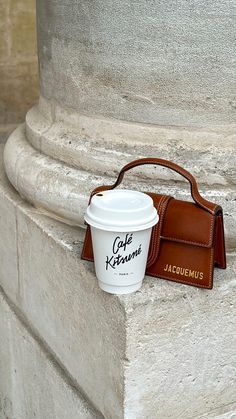 a cup of coffee sitting on top of a cement pillar next to a brown purse