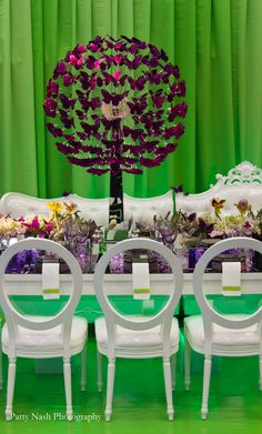 the table is set with white chairs and purple flowers in front of a green backdrop