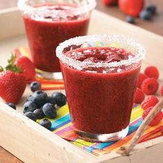 two glasses filled with fruit on top of a wooden tray next to strawberries and blueberries