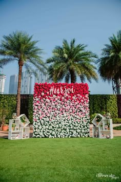 an outdoor wedding setup with flowers on the wall and white chairs in front of palm trees