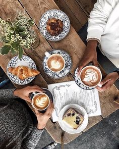 two people holding cups of coffee while sitting at a table with cakes and pastries