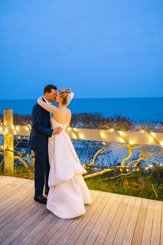 a bride and groom kissing on a deck with lights strung over the ocean in the background