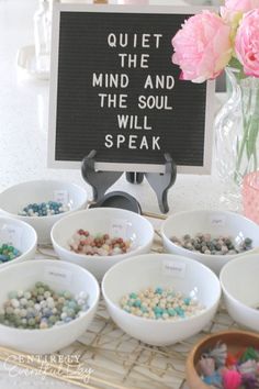 a table topped with white bowls filled with different types of beads next to a sign that says quiet the mind and the soul will speak