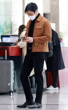 a man standing in an airport looking at his cell phone while wearing a face mask