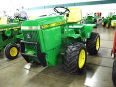 several green tractors are lined up in a warehouse with people standing around and looking at them