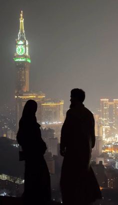 two people standing in front of a city skyline at night with a clock tower in the background