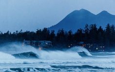 waves crashing on the beach in front of houses and mountains at night with lights lit up